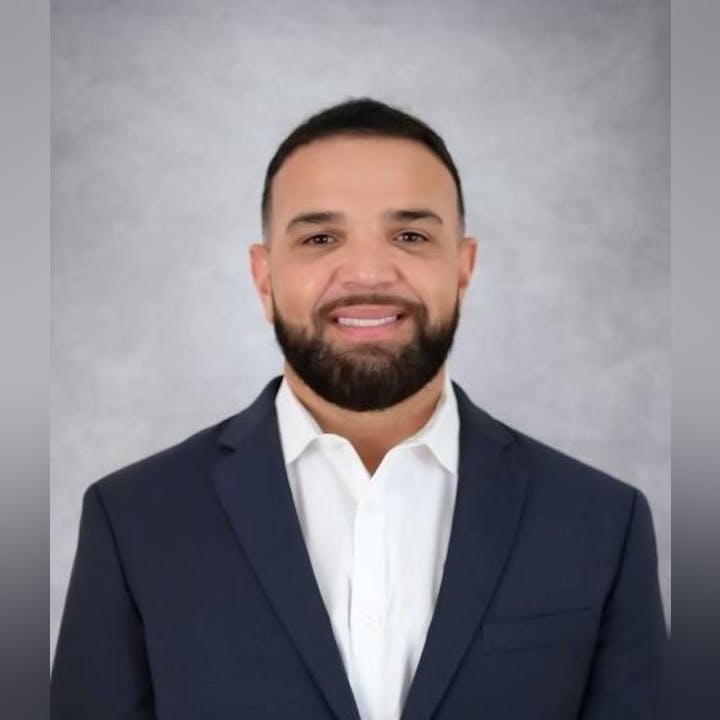 A professional-looking man with a beard smiles while wearing a suit and white shirt against a grey background.