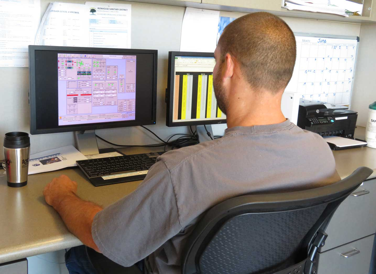 A person is sitting at a desk with two monitors displaying control systems and data, with a coffee cup nearby.