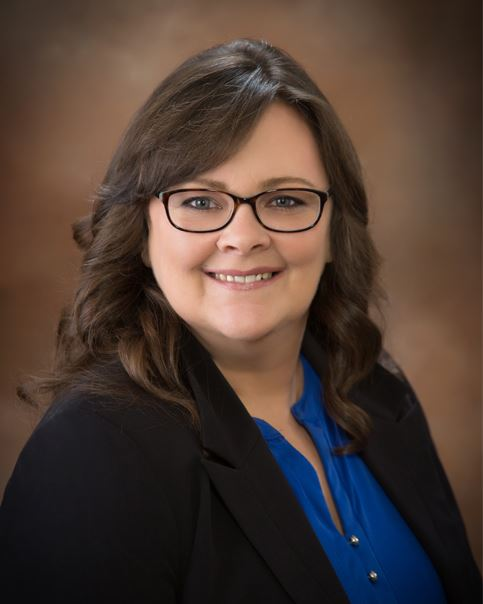 A smiling woman with brown hair, wearing glasses and a black blazer over a blue shirt, posing in front of a blurred background.