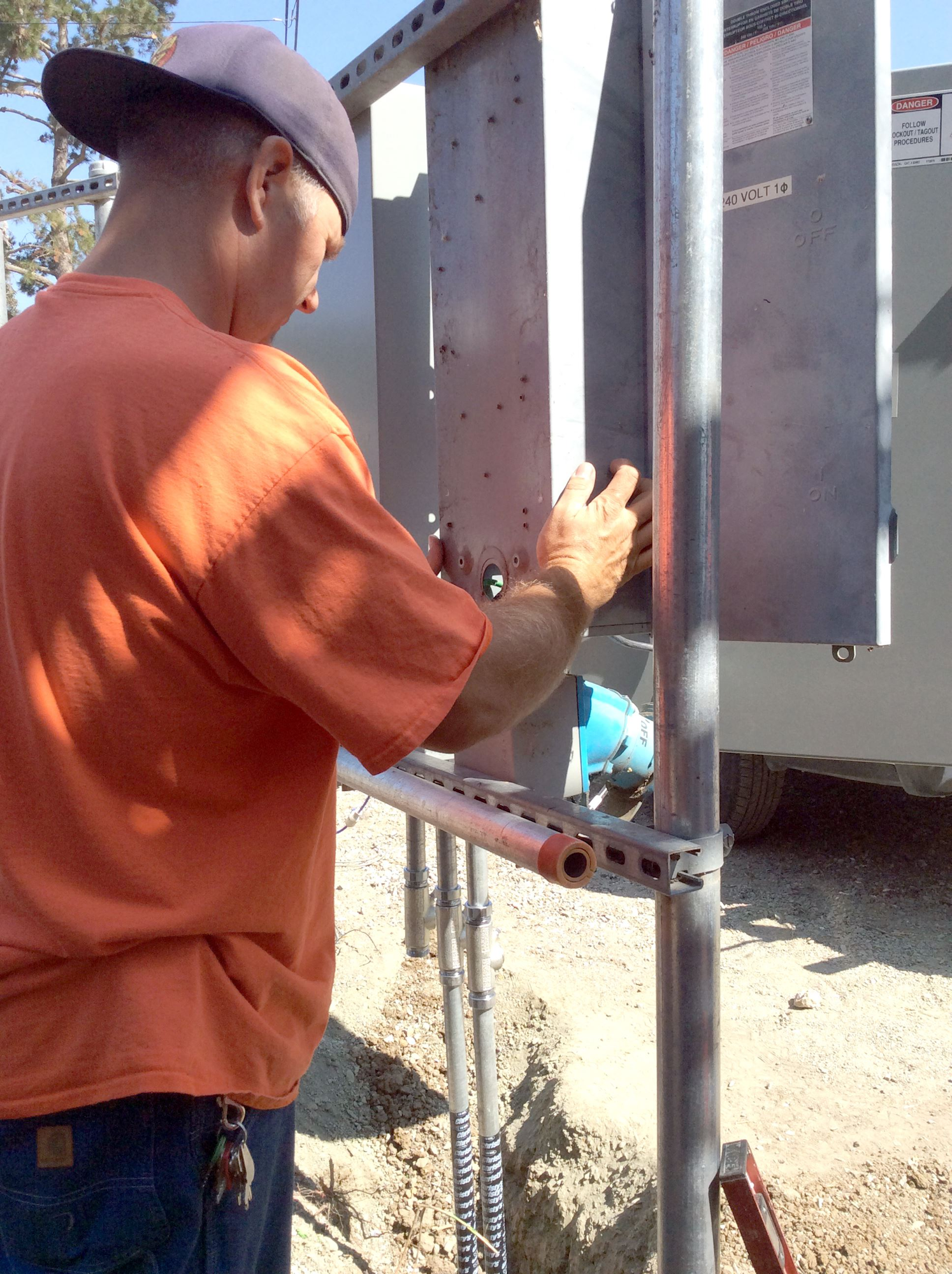 A person in an orange shirt is working on a metal panel, possibly related to electrical installation, outdoors in daylight.