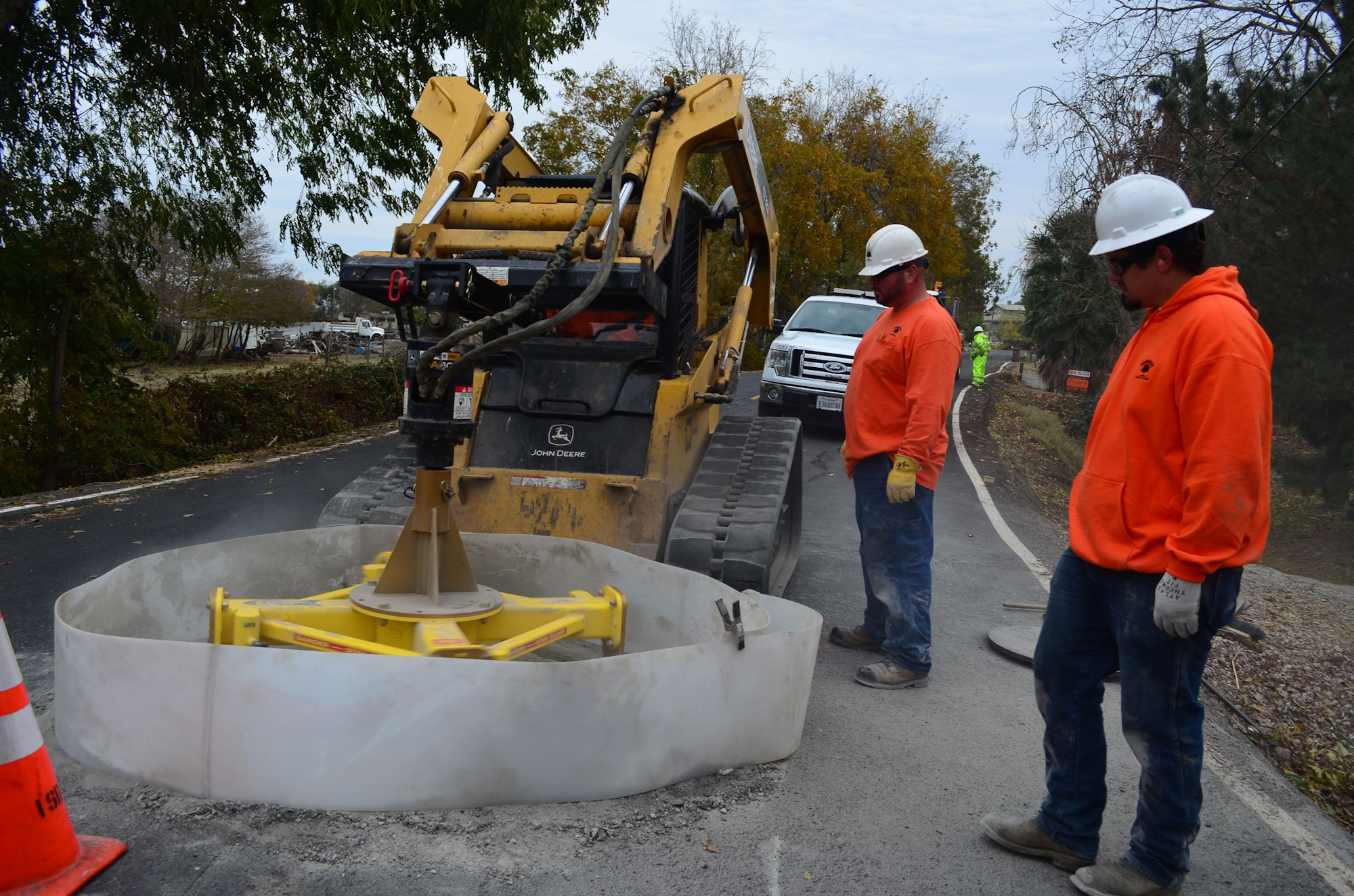 Construction workers operate machinery on a road, with a focus on a skid steer loader and a circular cutting attachment.