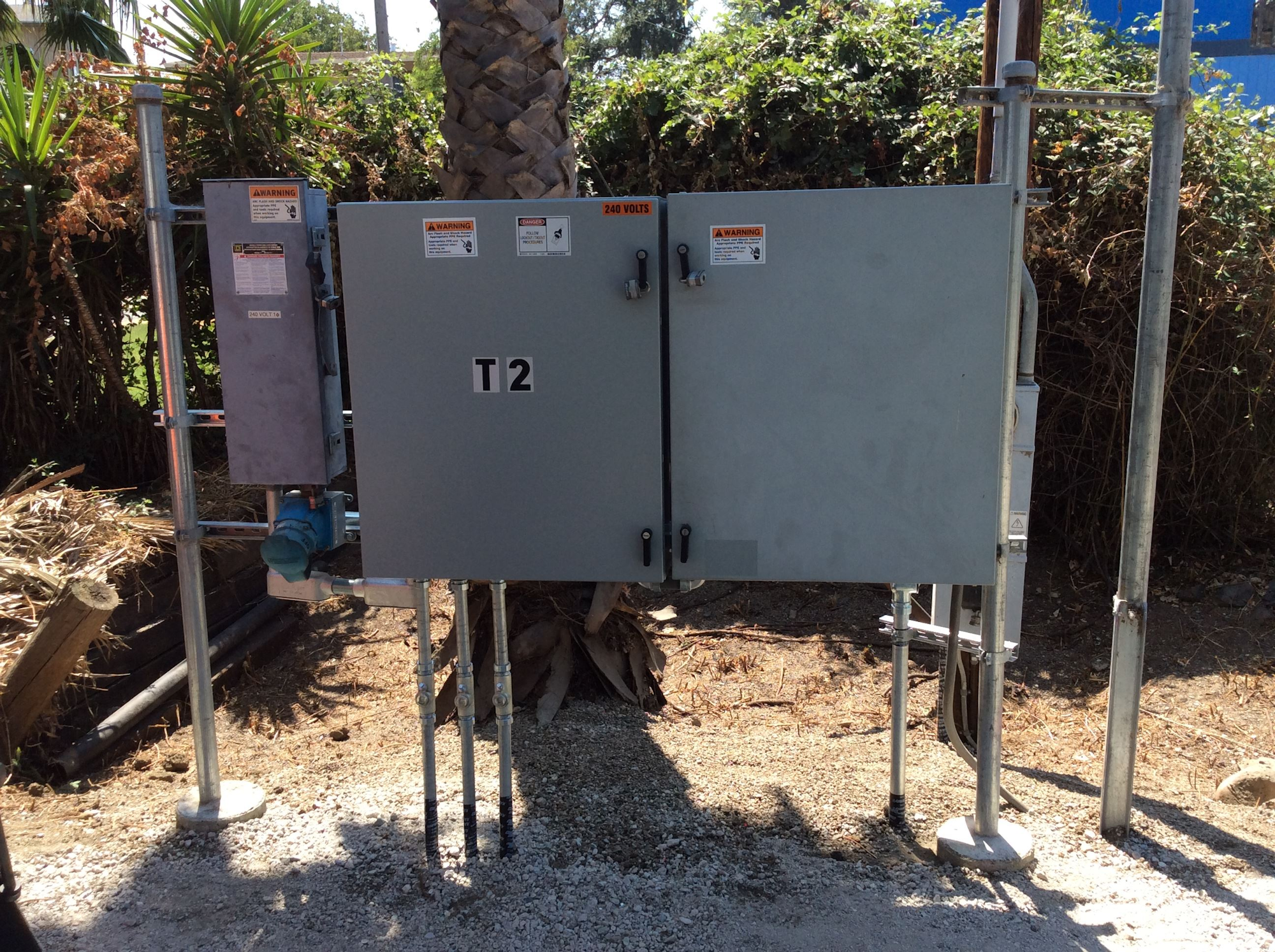 The image shows electrical equipment with metal enclosures, labeled "T2" and warning signs, set in a landscaped area.