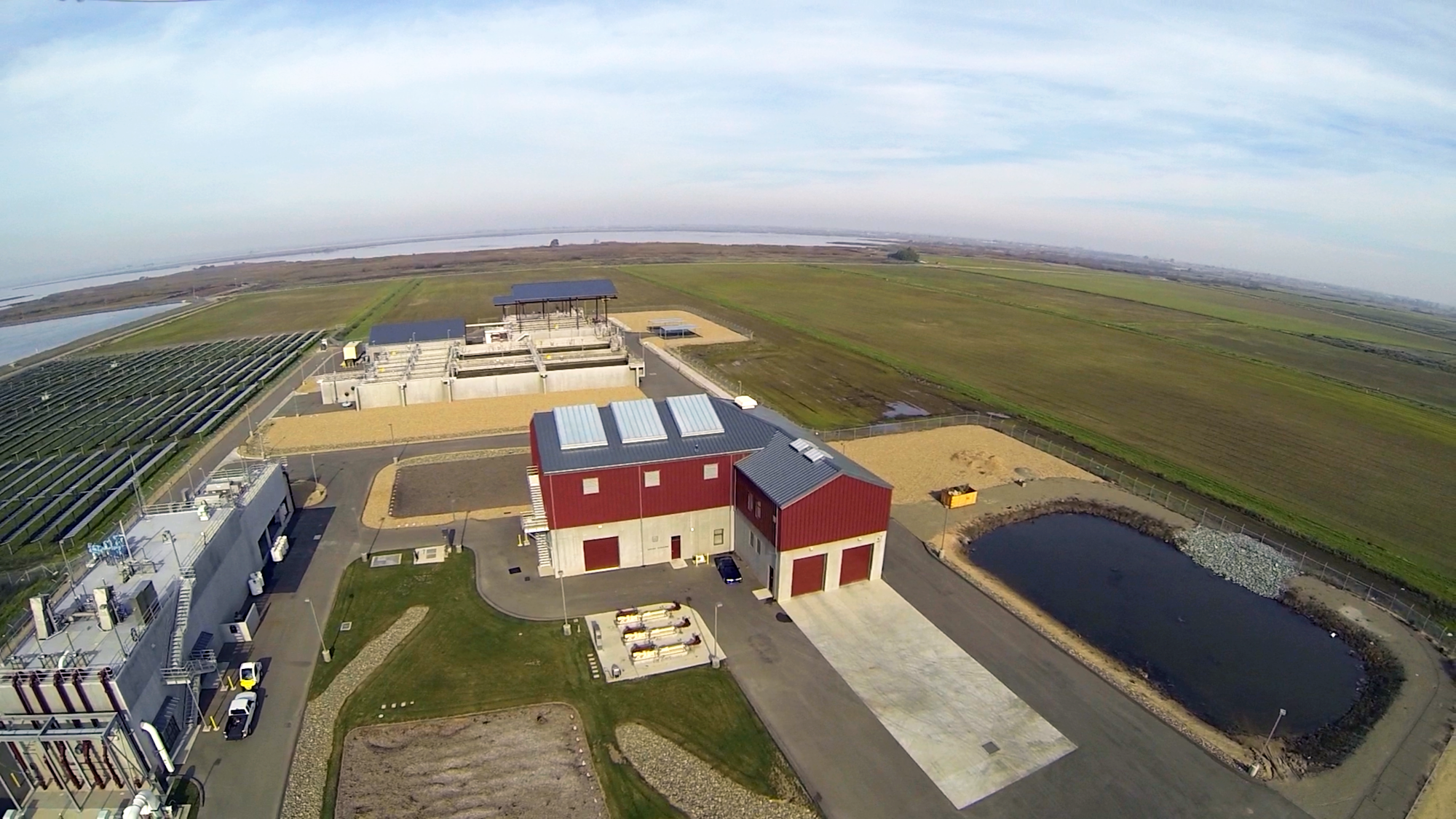An aerial view of a facility with several buildings, solar panels, fields, and a small pond in a rural setting.