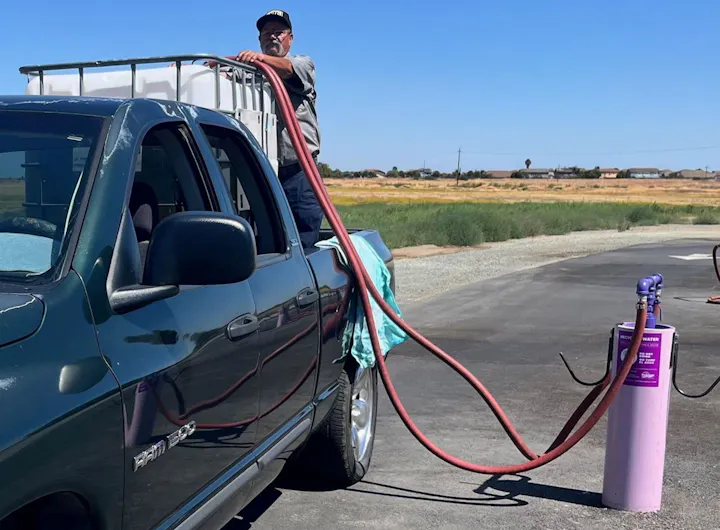 An older man stands on a truck, using a hose to fill a container from a purple water dispenser in a rural setting.