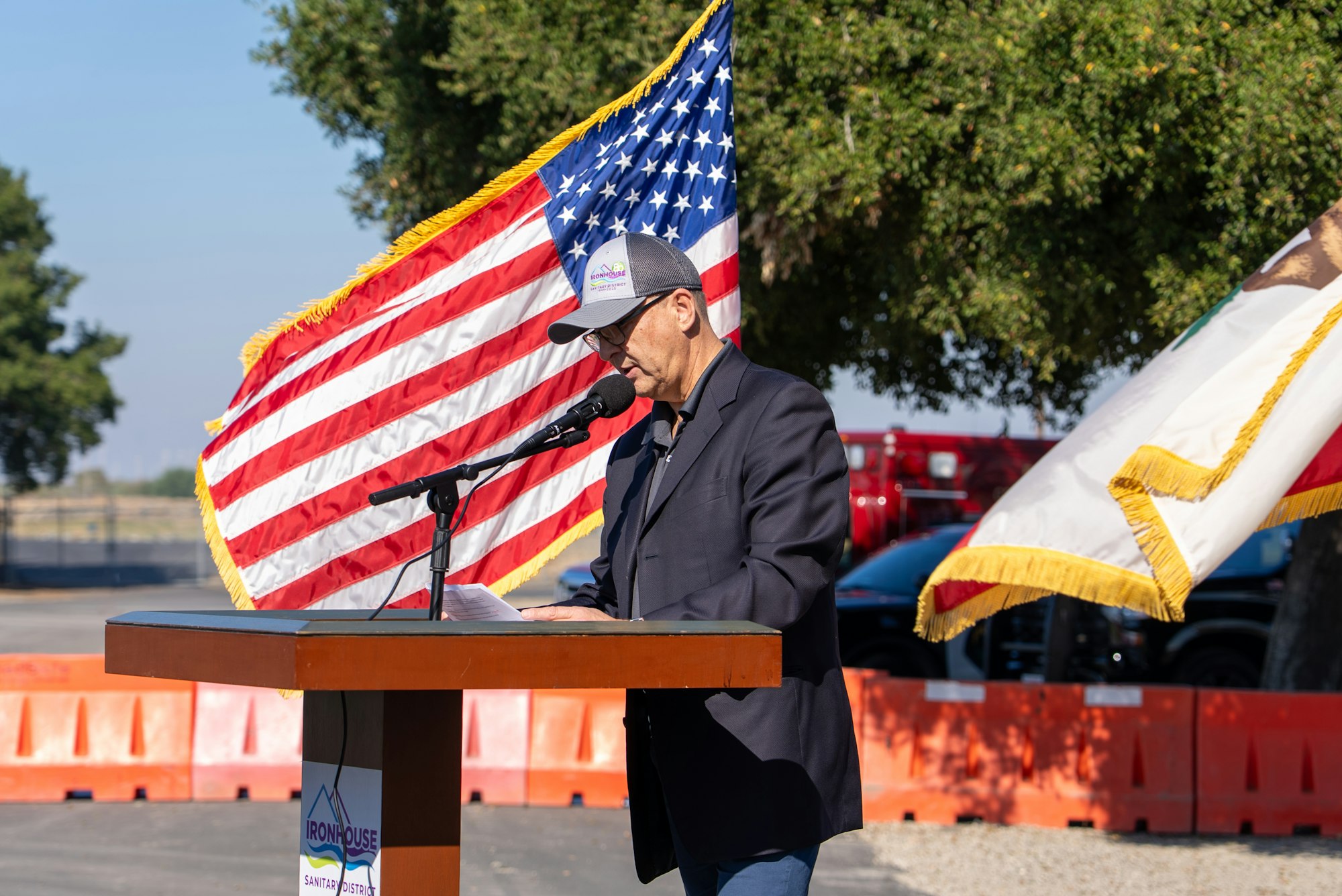 A speaker addresses an audience at an outdoor event, with American and California flags in the background.
