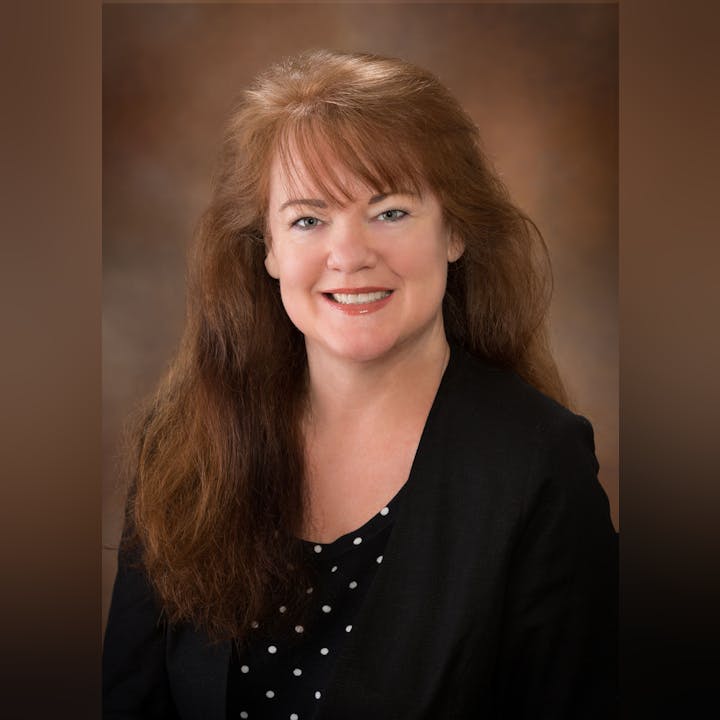 A woman with long, wavy reddish-brown hair and a black blazer smiles for a professional headshot. Background is softly blurred.