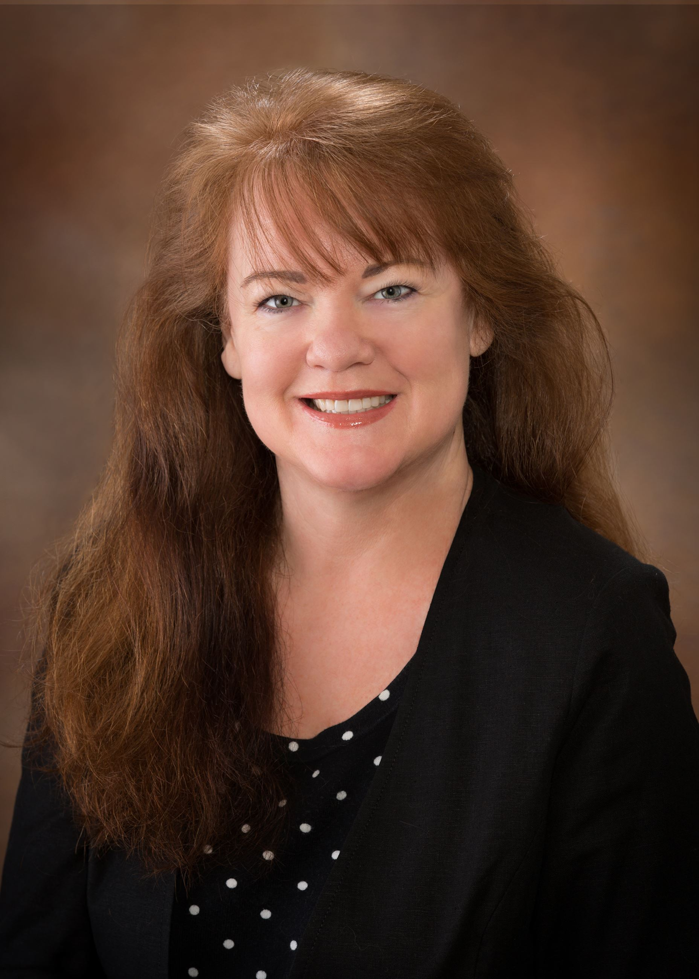 A woman with long, wavy reddish-brown hair and a black blazer smiles for a professional headshot. Background is softly blurred.