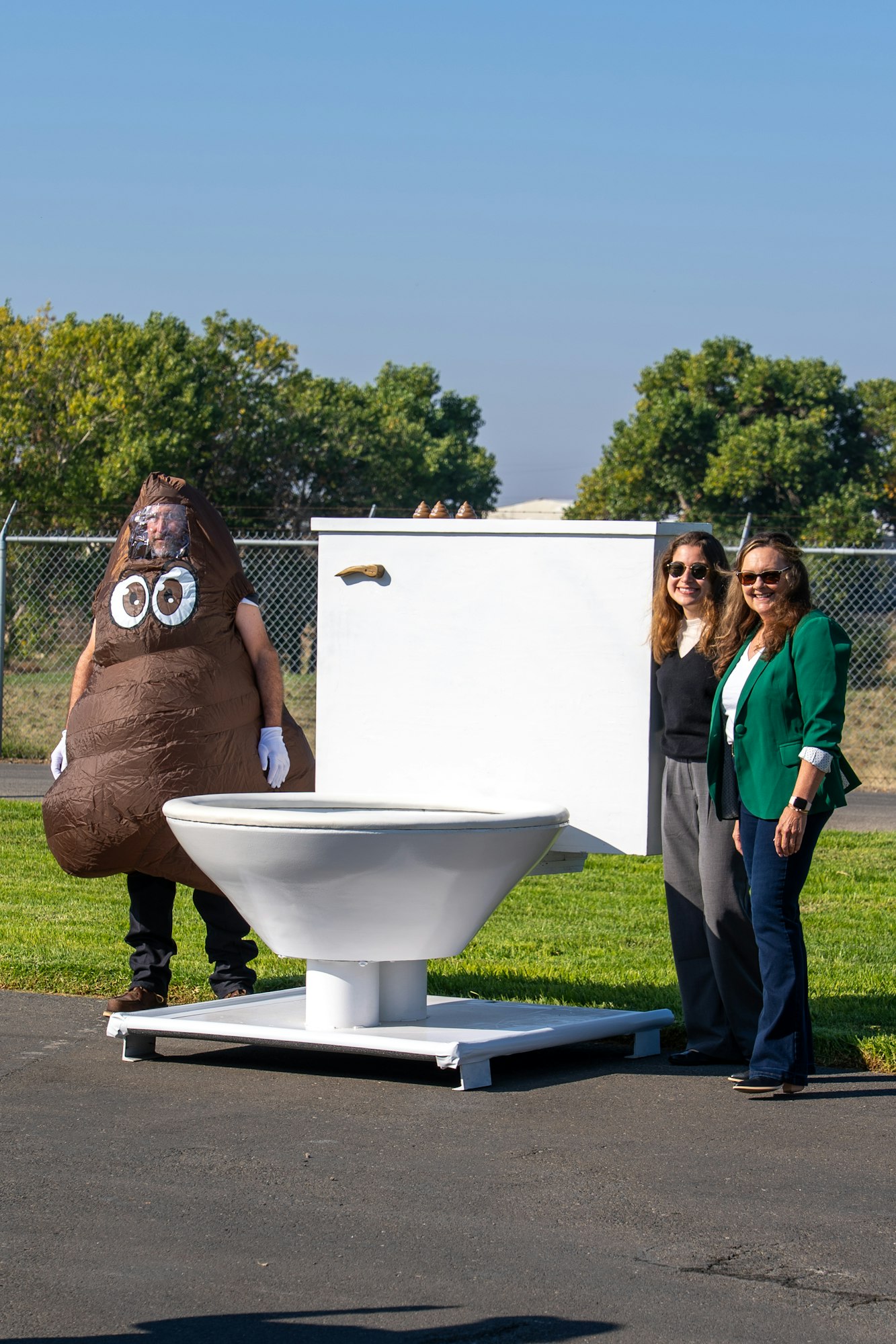 A person in a poop costume poses with two women near a large toilet and a blank sign in a grassy area.