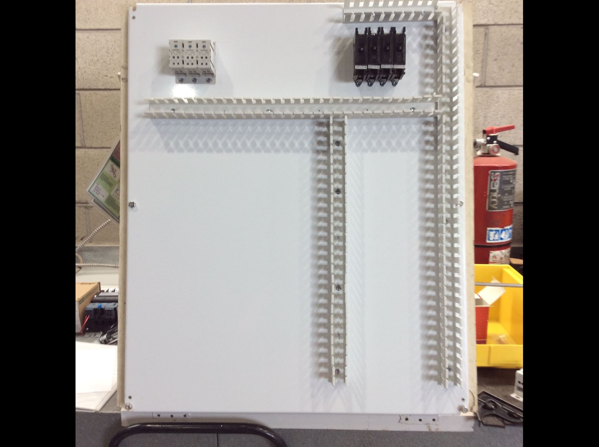 The image shows a white panel with mounting tracks, some electrical components, and a fire extinguisher in the background.