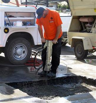 A worker using a jackhammer to break concrete at a construction site, with equipment nearby.