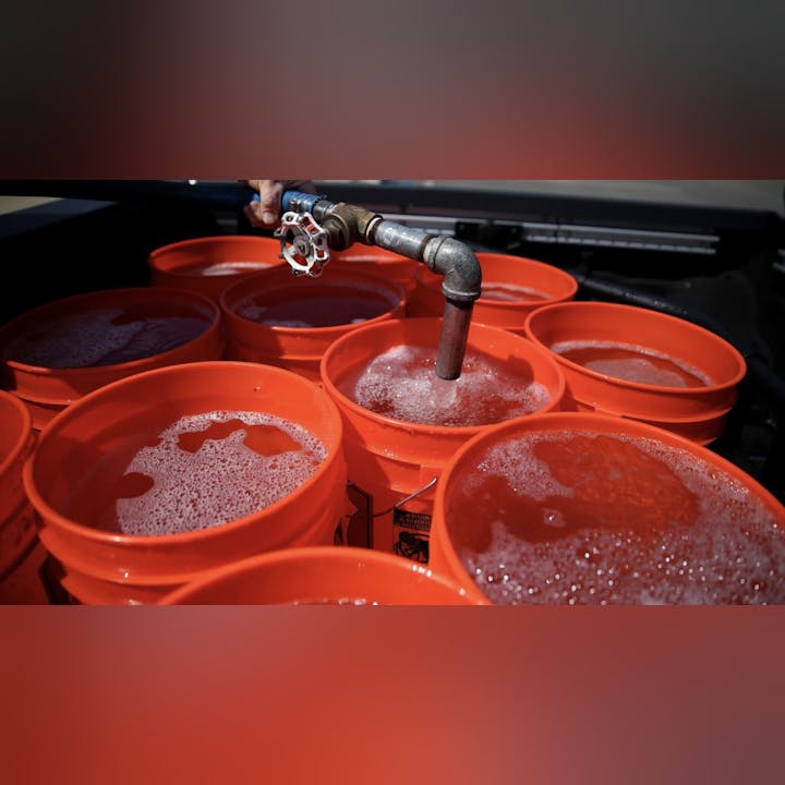 The image shows orange buckets being filled with water or liquid from a spout, creating bubbles on the surface.