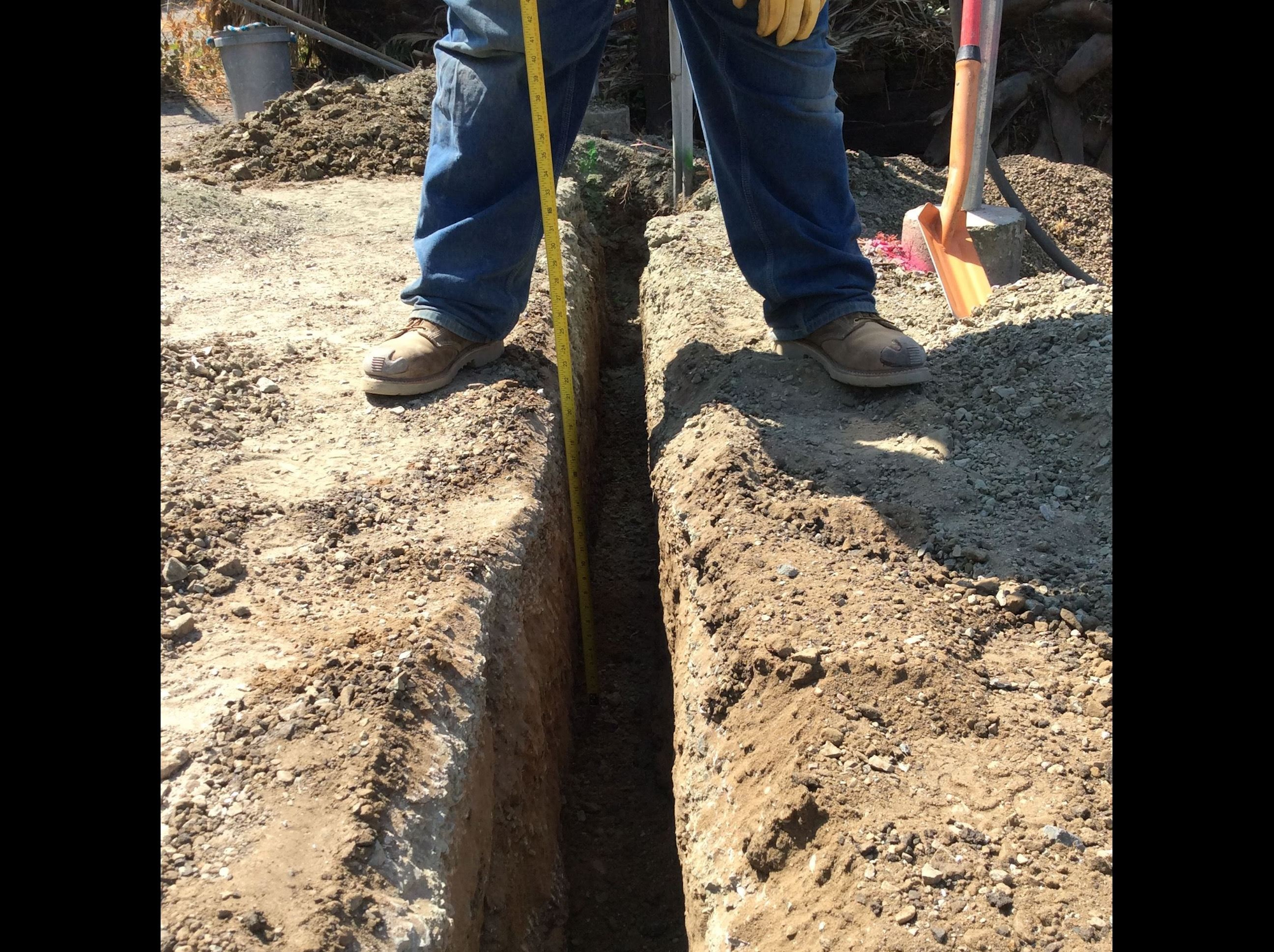 A person stands over a deep trench, measuring its depth with a tape measure, surrounded by gravel and dirt.