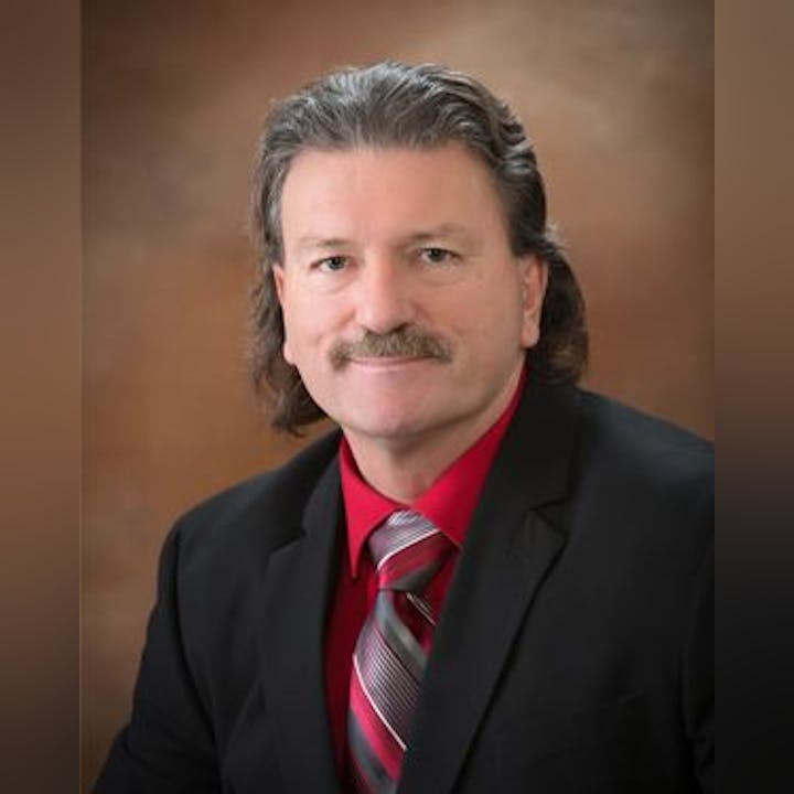 A man with long hair and a mustache, dressed in a black suit and red shirt, poses for a professional photo.