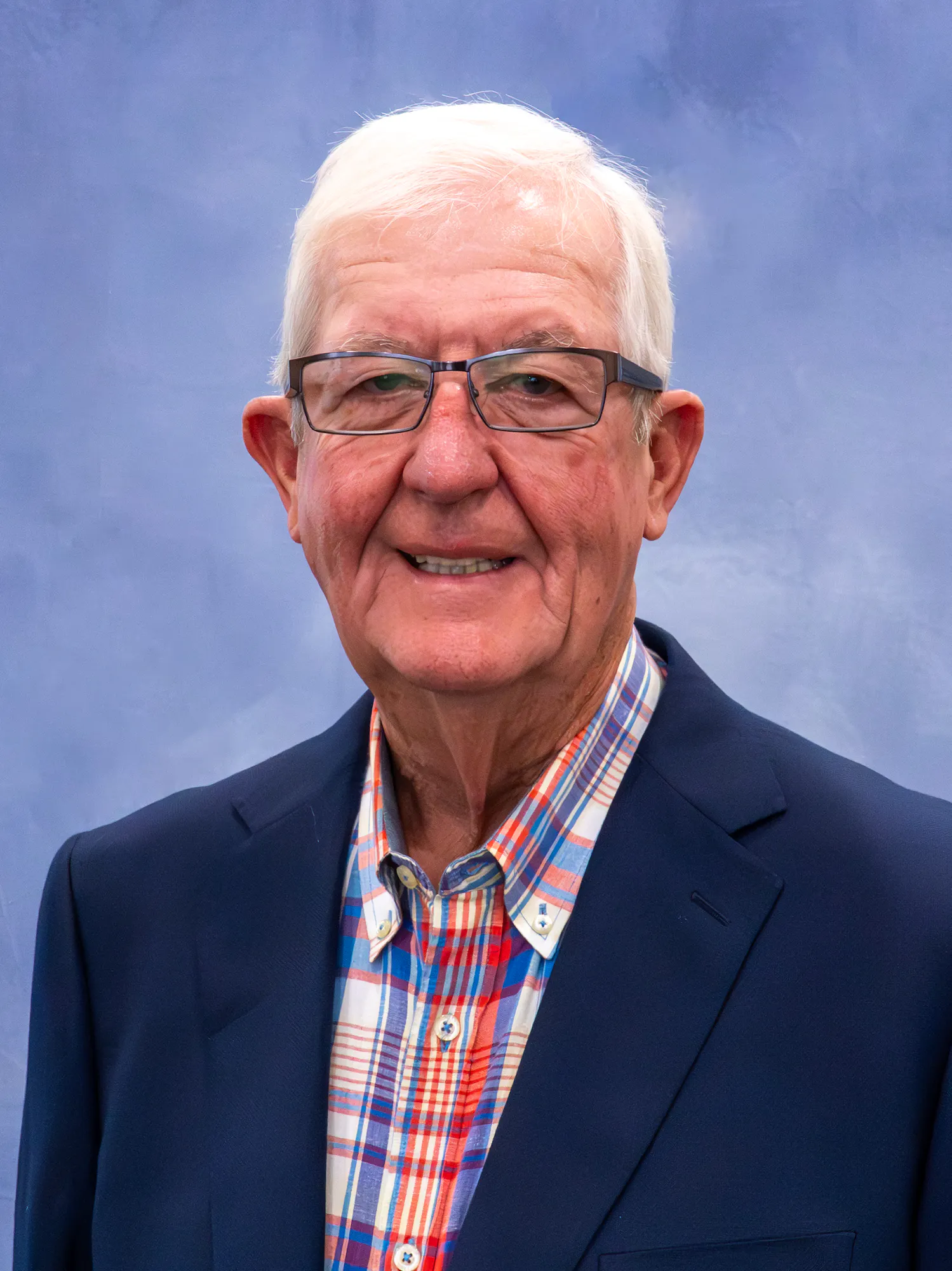 The image features an elderly man with short white hair, glasses, and a colorful plaid shirt, smiling against a blue background.