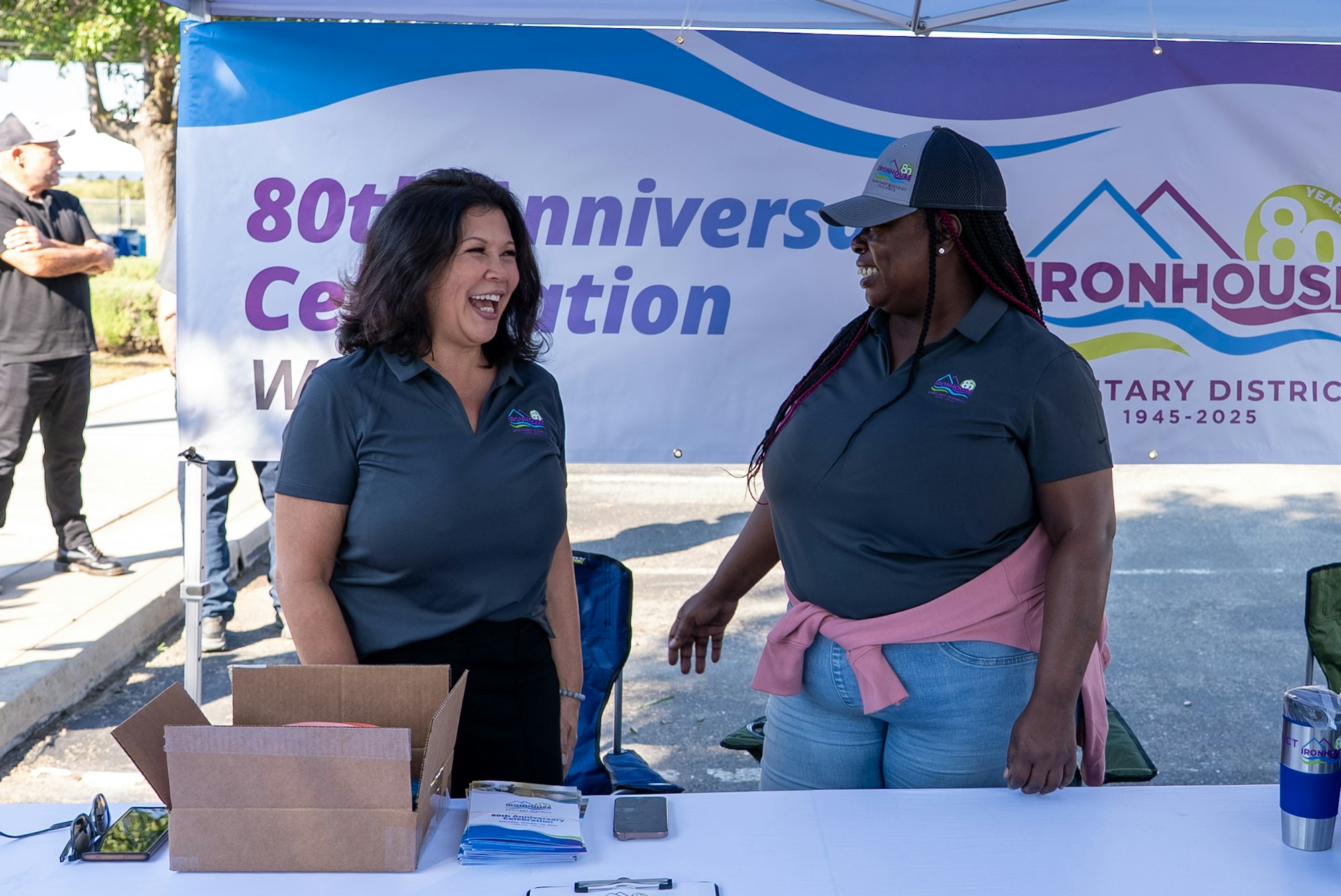 Two women are smiling and chatting at a booth celebrating the 80th anniversary of a district. A banner is visible behind them.