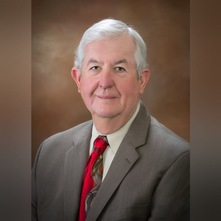 A man in a suit with a patterned tie poses for a professional portrait against a neutral background.