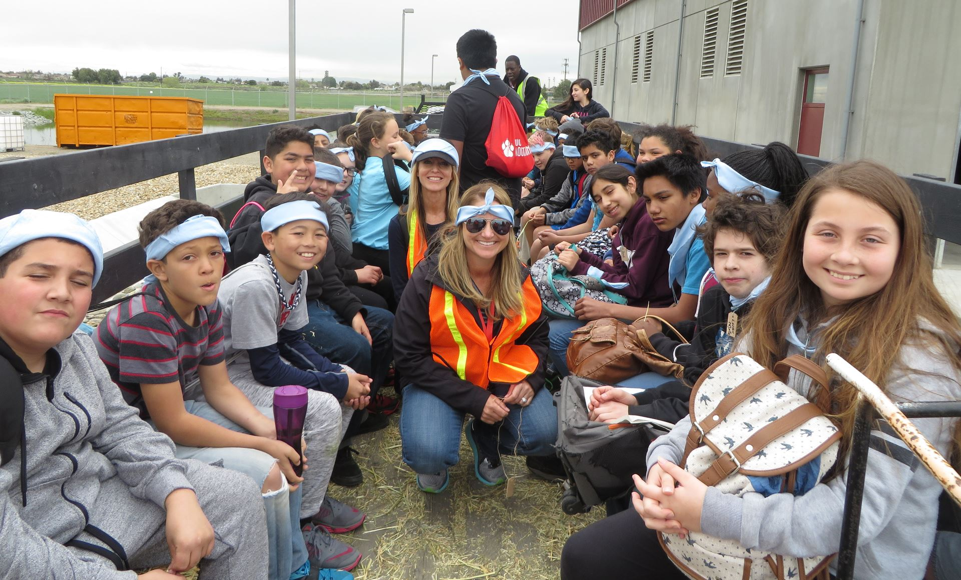A group of students and adults, some wearing blue headbands, sit together in a casual outdoor setting, seemingly on a field trip.