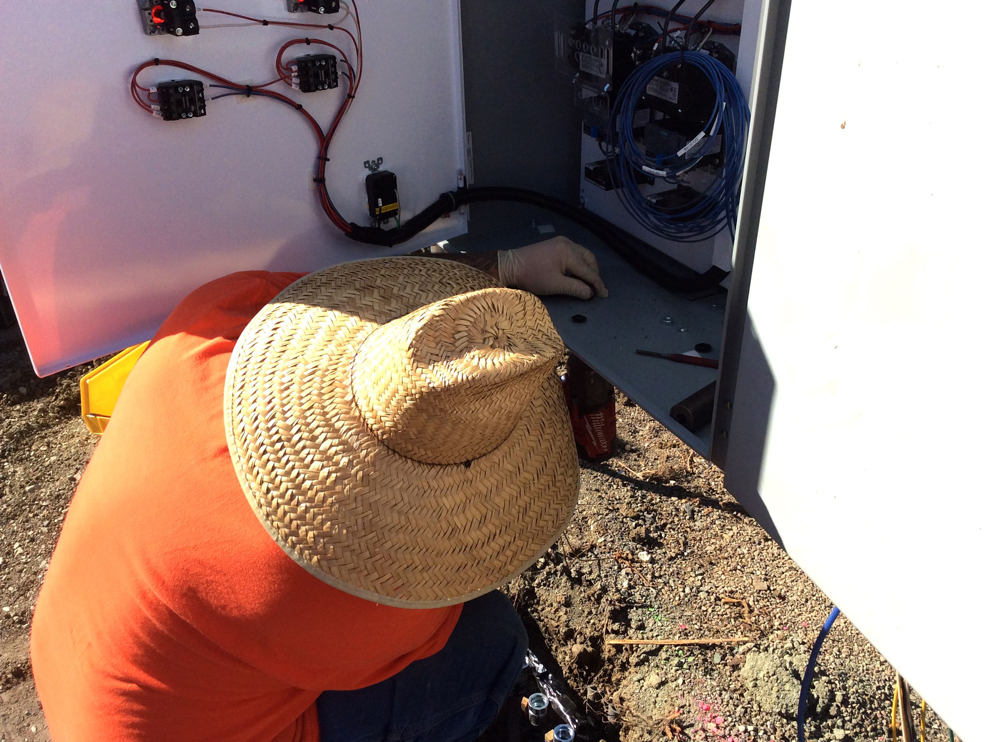 A person wearing a straw hat works on electrical wiring inside a cabinet, surrounded by tools and dirt.