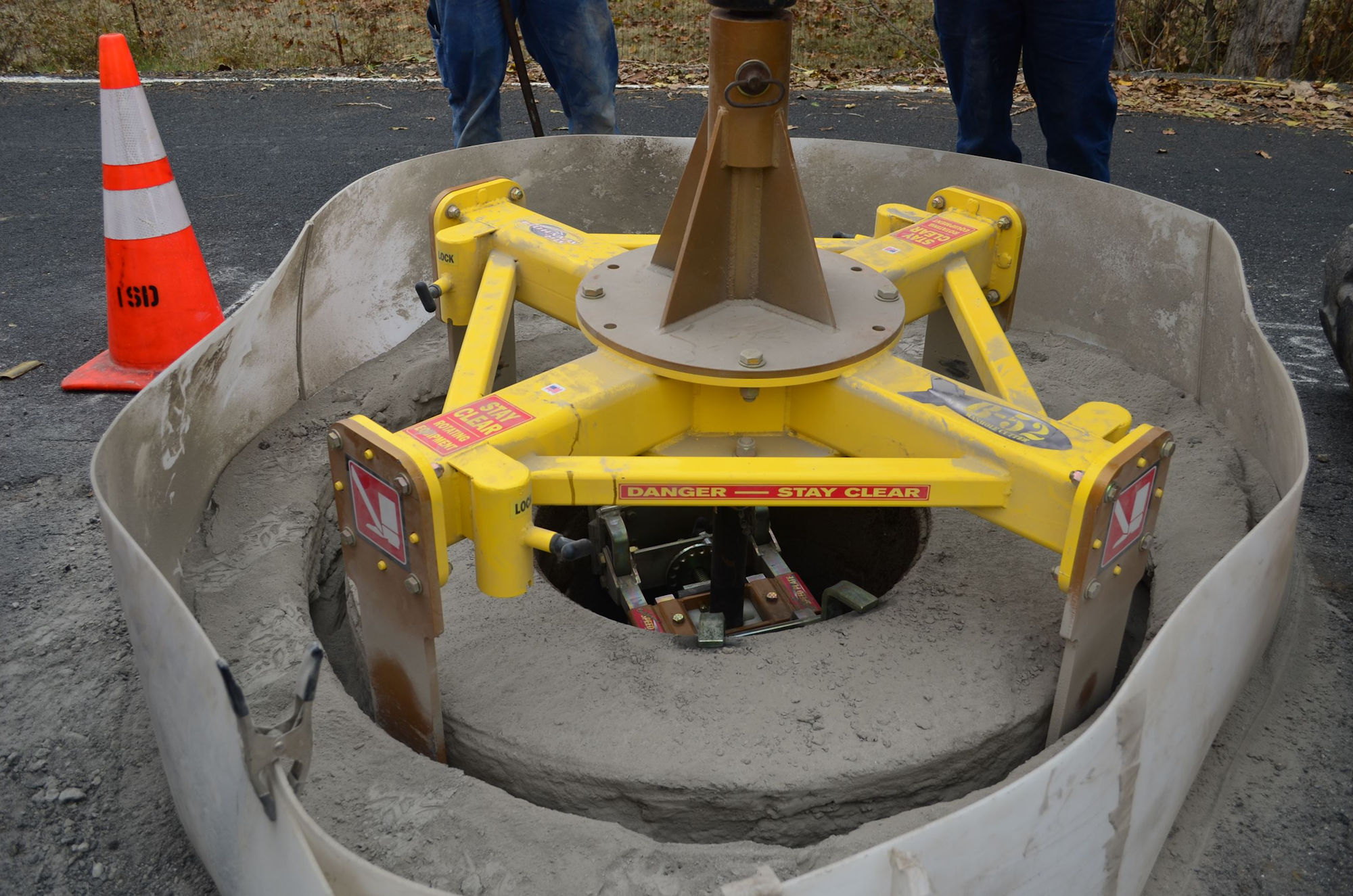 The image shows a yellow construction tool for mixing concrete, surrounded by a safety barrier and a warning cone.