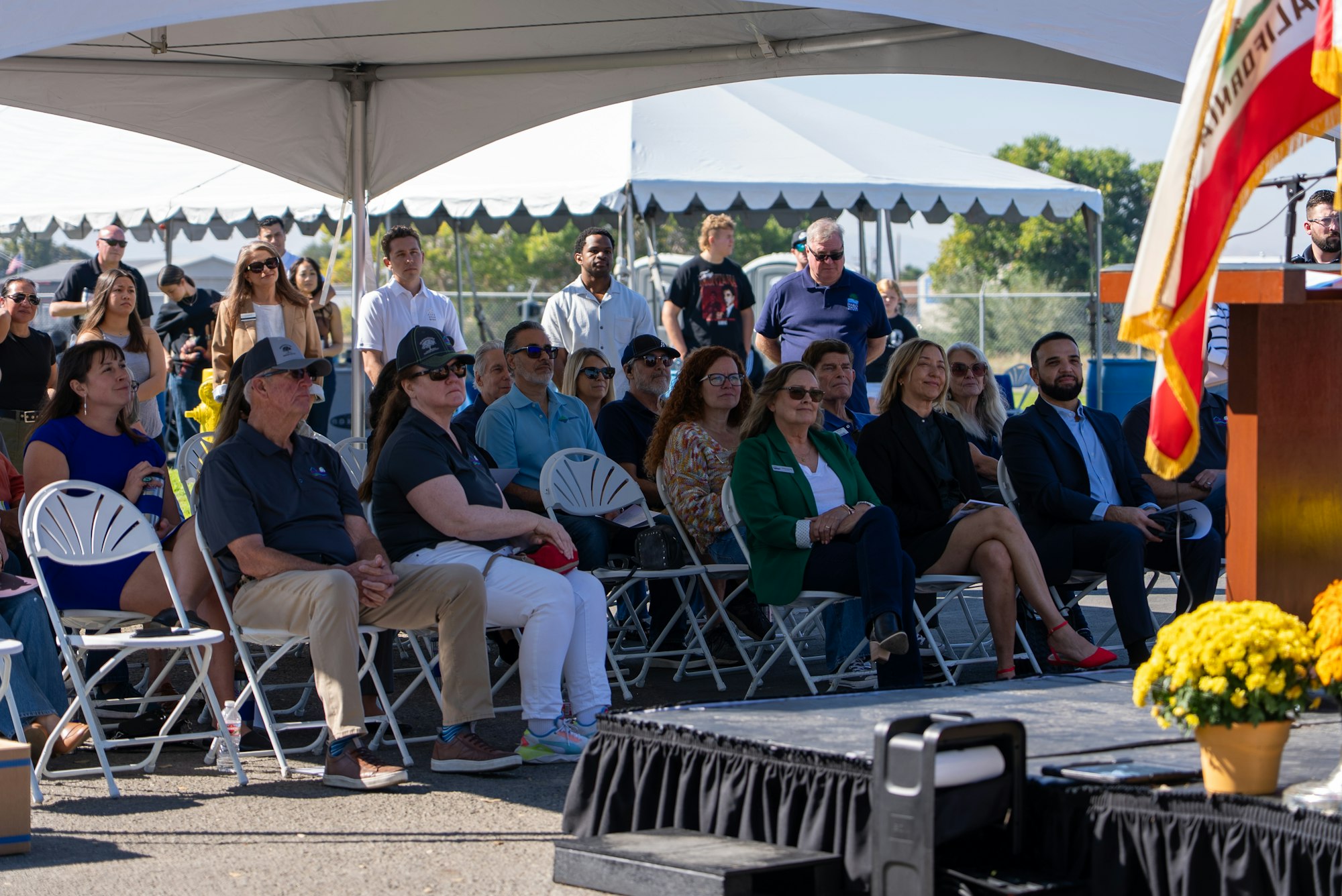 A diverse group of people seated under a tent, listening to a speaker at an outdoor event with chairs and a podium.