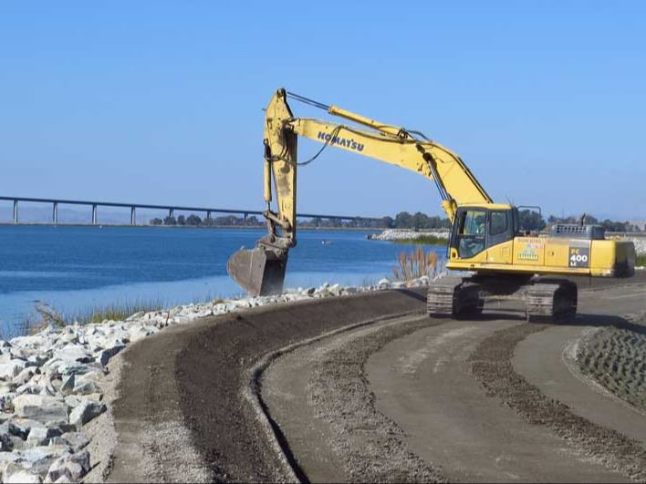An excavator is working near a body of water, shaping the shoreline and preparing the area for development.