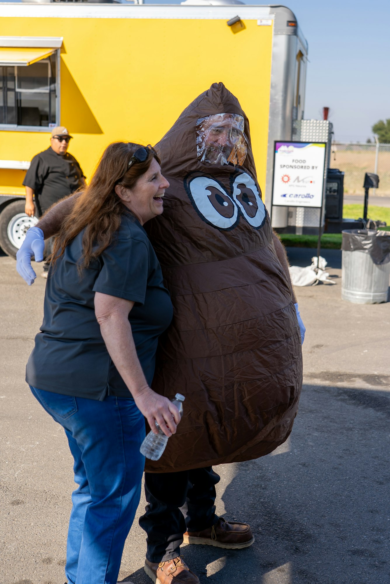 A woman laughs with someone in a large brown poop costume at an outdoor event, with food trucks in the background.