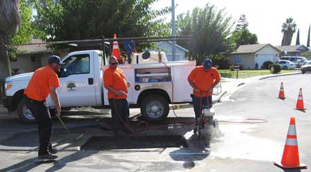 Workers in orange shirts repair a road, using equipment near a utility truck, with traffic cones marking the work area.
