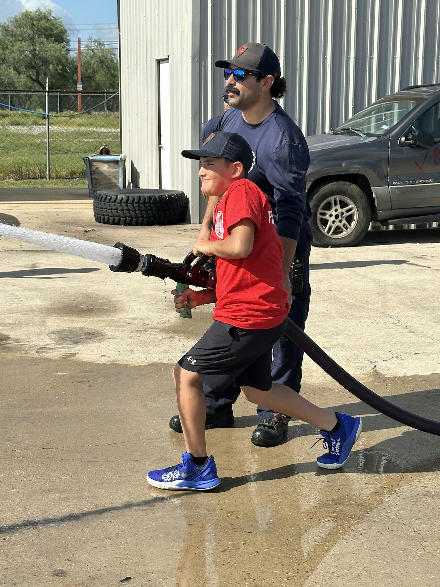 A child and an adult using a fire hose outdoors, with a vehicle and building in the background.