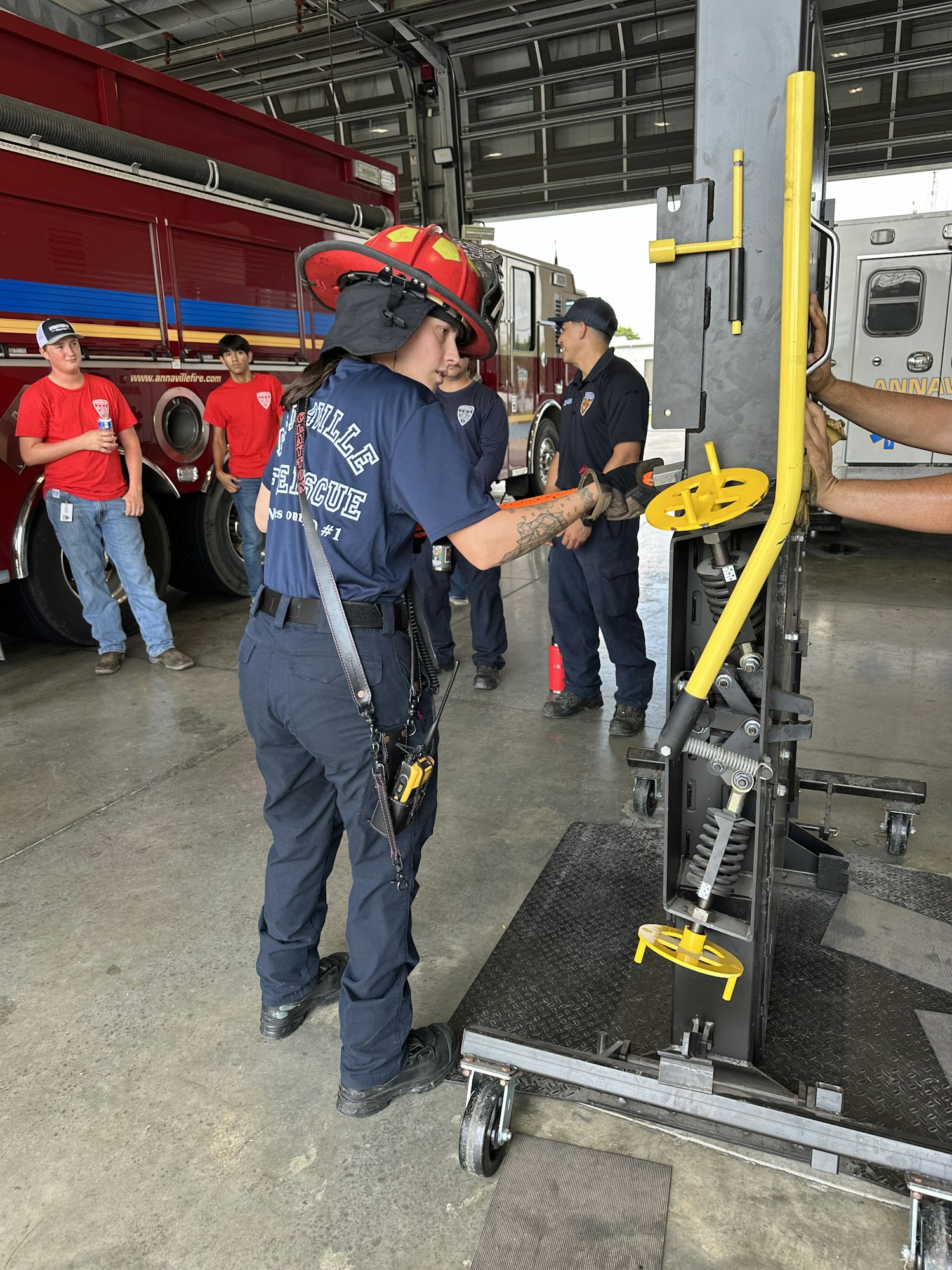 Firefighters in uniform, one with a red helmet, around equipment in a fire station, preparing or inspecting machinery.