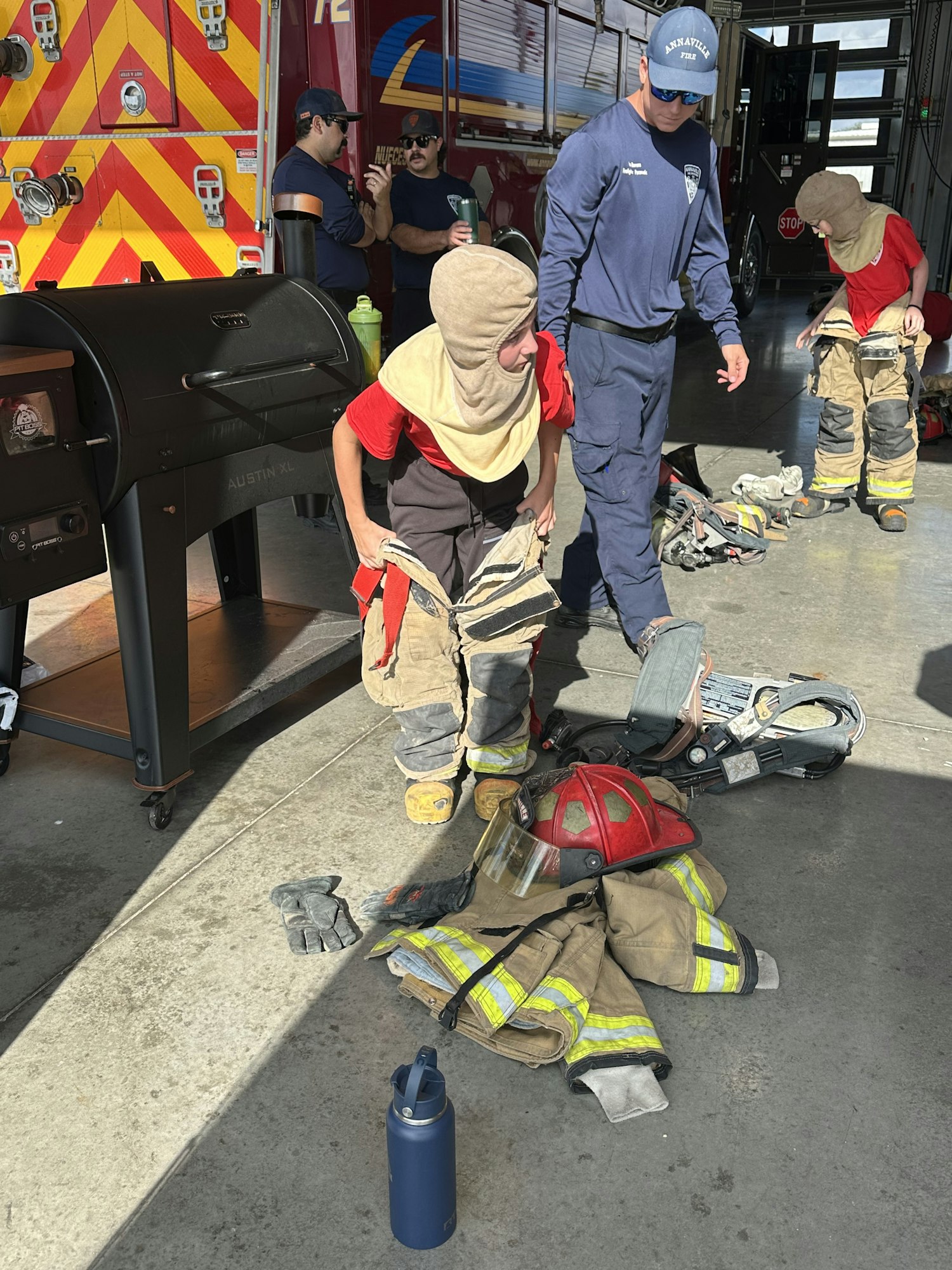 People in a fire station, wearing and handling firefighting gear, with a fire truck and equipment in the background.