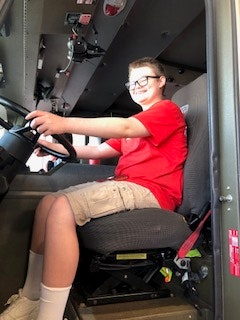 A person in a red shirt and shorts is sitting in the driver's seat of a vehicle, holding the steering wheel.