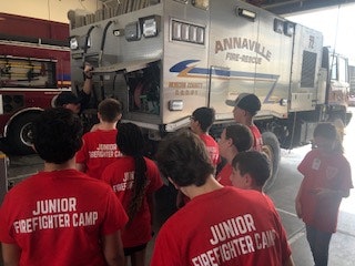 A group of kids in red "Junior Firefighter Camp" shirts stands near a fire rescue vehicle inside a fire station.