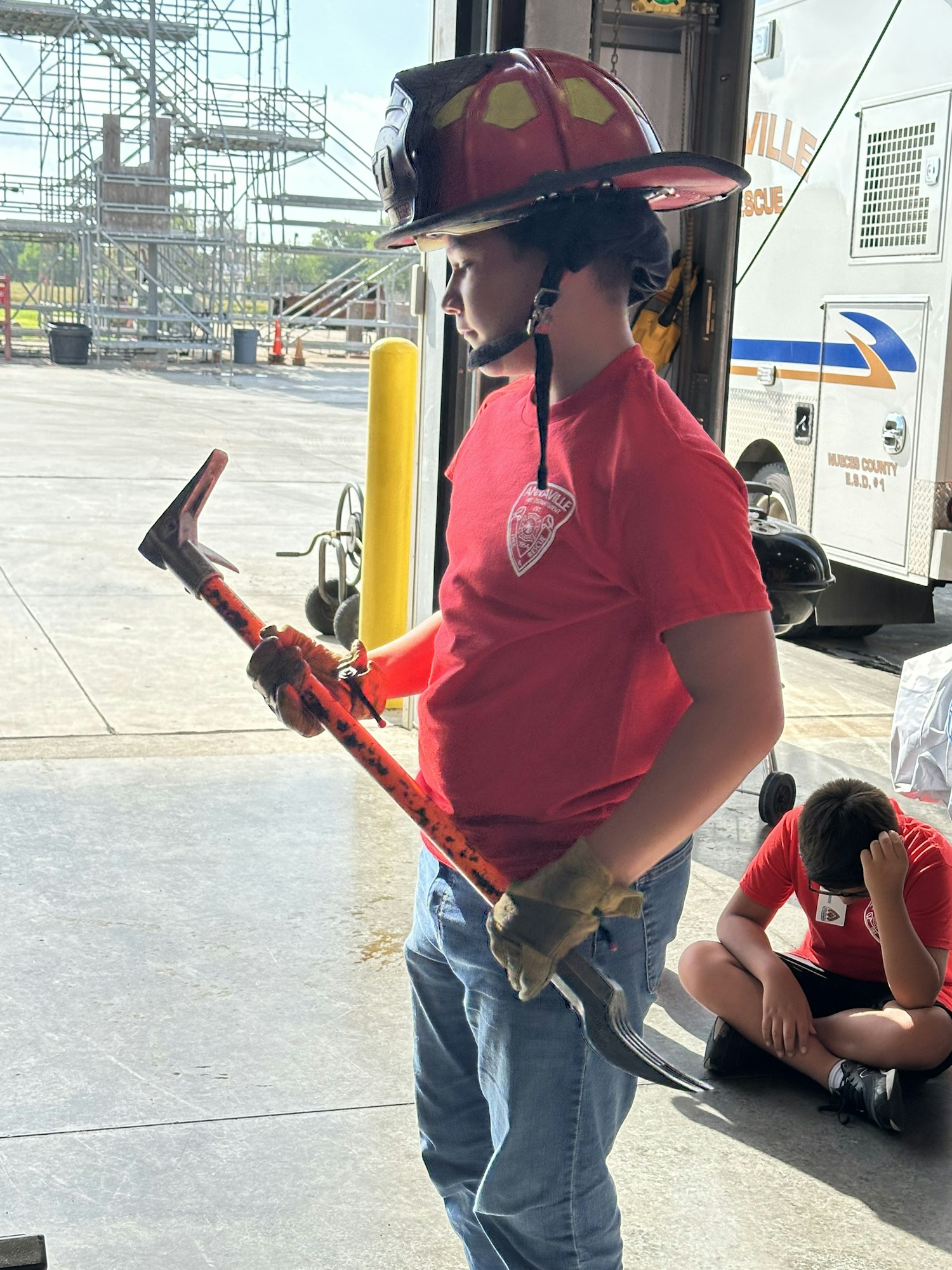 A person in a firefighter helmet holds a tool outside a garage near an emergency vehicle; another person sits on the floor.