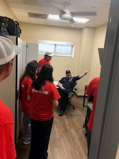 People in red shirts in an office room, with one person sitting, appearing to give instructions or directions.