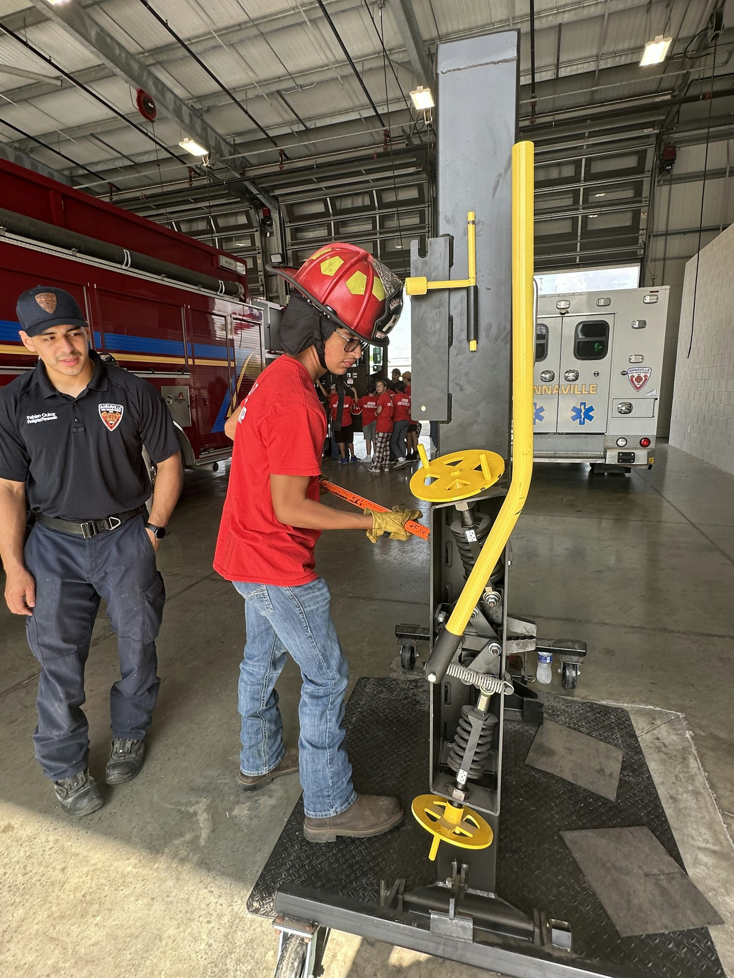A person in a red helmet and shirt operates a mechanical device, while another stands nearby in a garage with emergency vehicles.