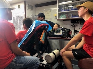 Four people inside an ambulance, one wearing a uniform showing equipment, others in casual clothes listening.