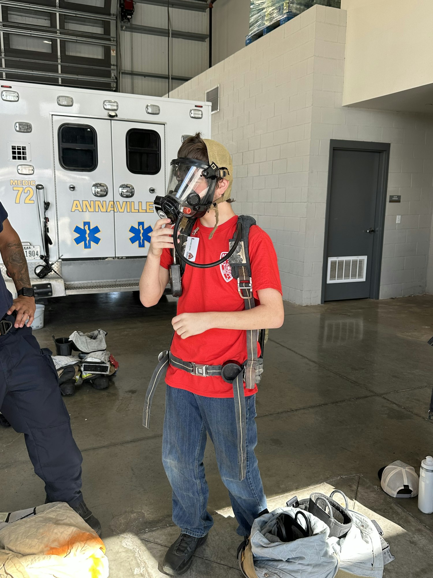 A person wearing a respirator and harness, standing by an ambulance in a garage.