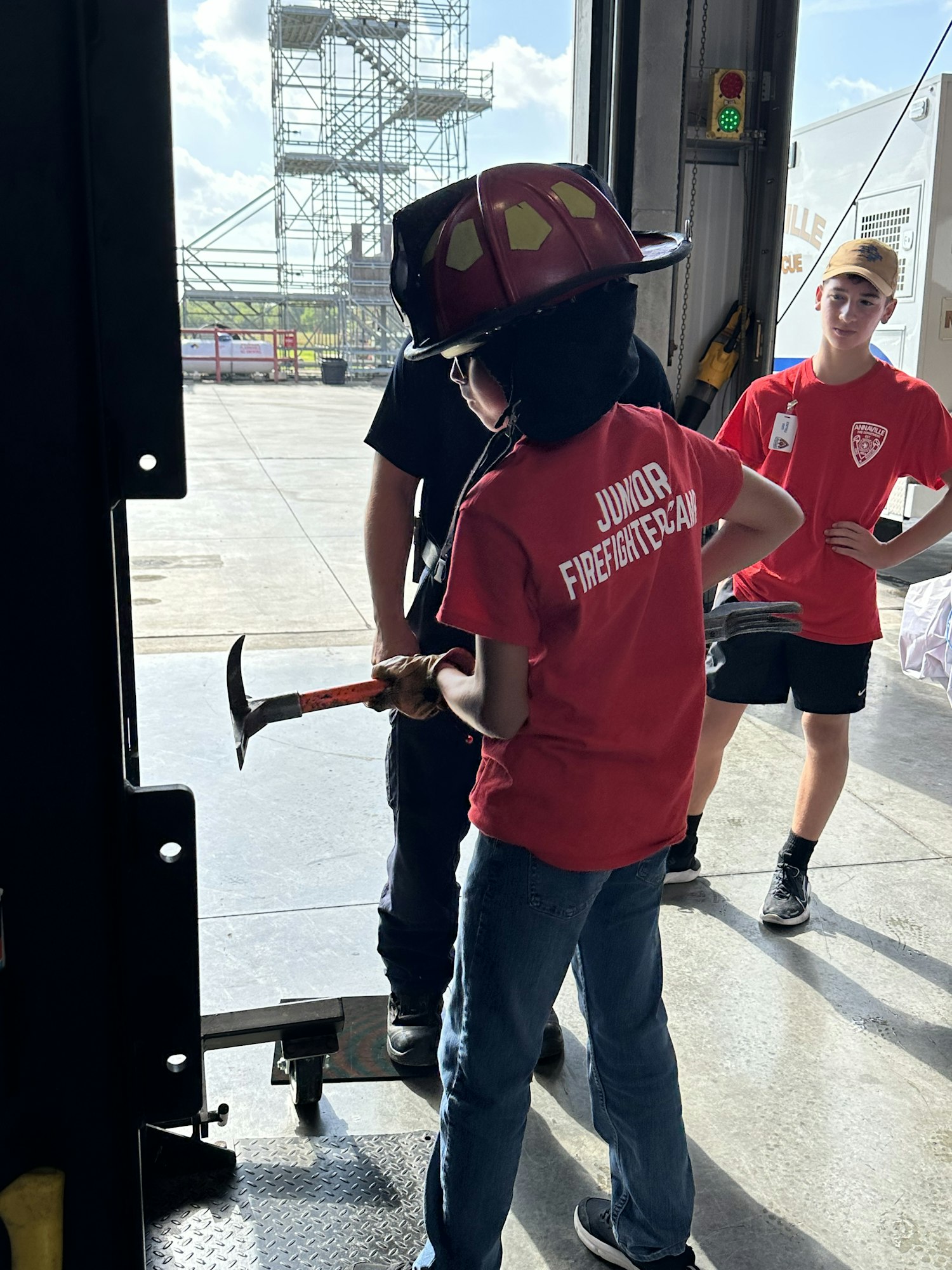 A child wearing a "Junior Firefighter" shirt and helmet holds a tool, with another person in the background.