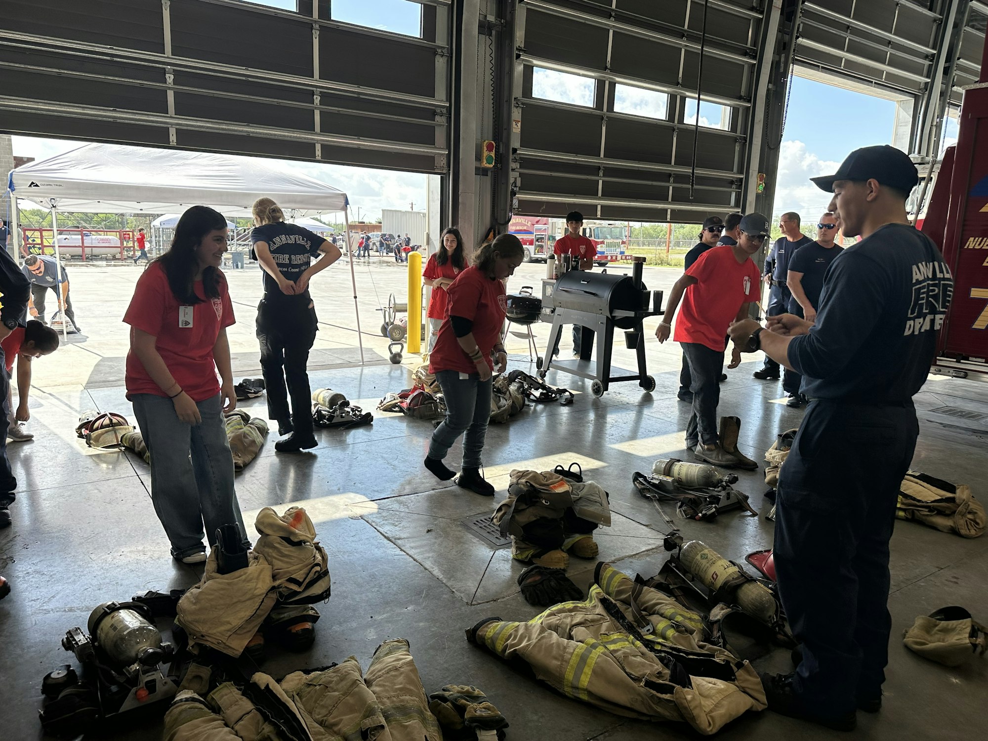 People in a fire station, some in red shirts and others in dark uniforms, surrounded by firefighting gear and equipment.