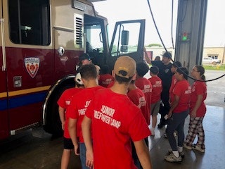 Group of kids in "Junior Firefighter Camp" shirts, gathered near a fire truck inside a fire station.