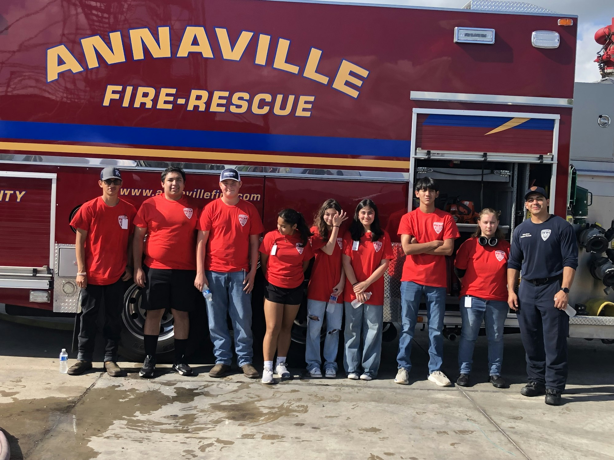 A group of people in red shirts stand in front of an "Annaville Fire-Rescue" vehicle, next to a firefighter.