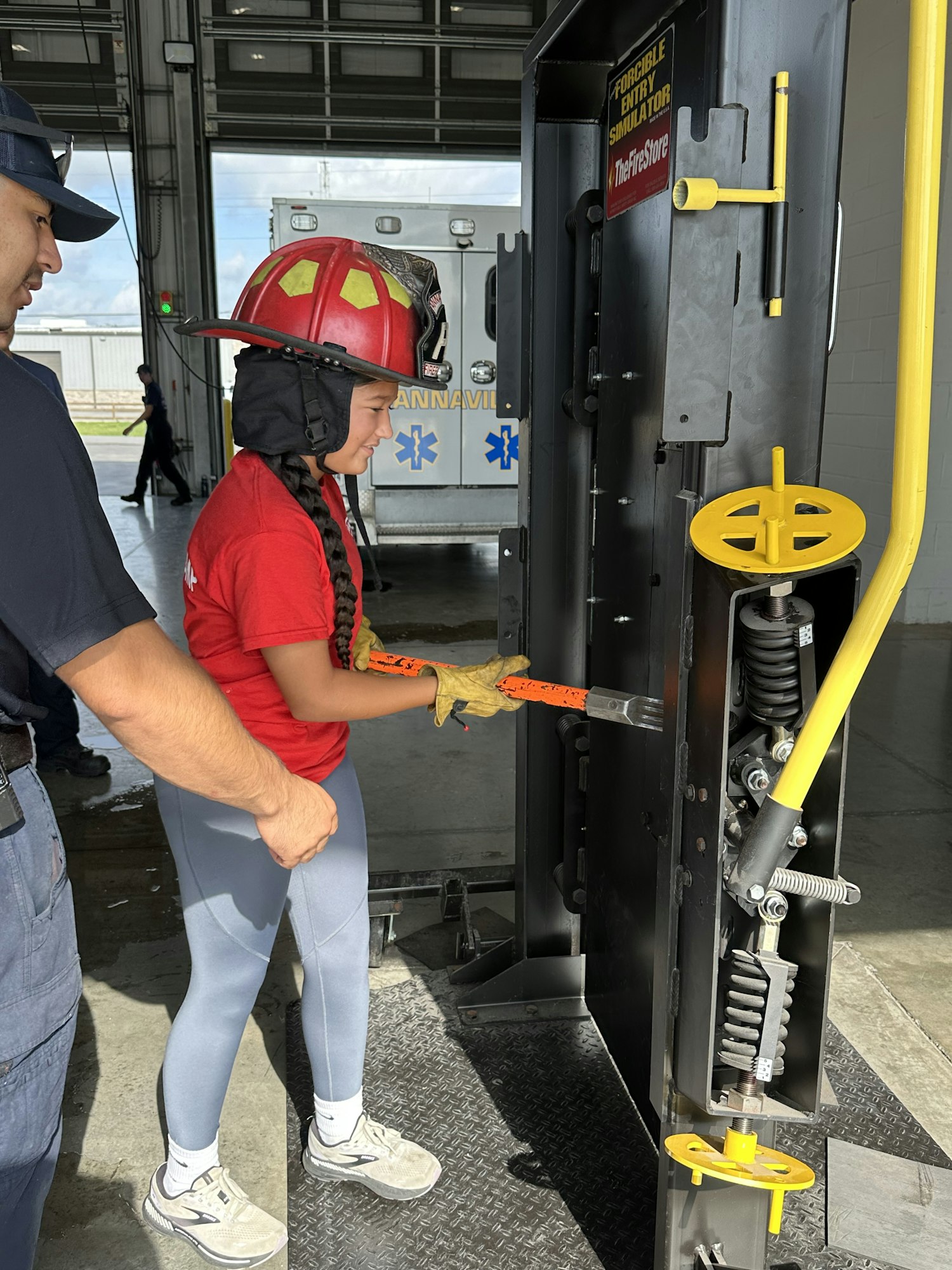 A person in firefighting gear operates rescue equipment with guidance from another individual in a garage setting.