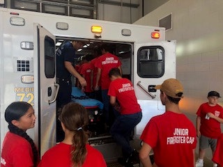 Kids in red "Junior Firefighter Camp" shirts gather around and inside an ambulance with a firefighter.