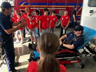 A firefighter demonstrates rescue equipment to children wearing red shirts during a tour at a fire station.