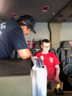 A person in a red shirt is seated inside a vehicle with a firefighter, who is wearing a navy uniform and hat, engaging in discussion.