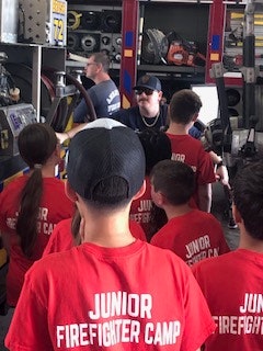 A group of people in red "Junior Firefighter Camp" shirts gather around a firefighter showing equipment.