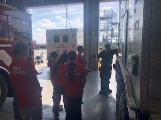 A group of kids in red shirts labeled "Junior Firefighter" listens to a firefighter near emergency vehicles in a garage.