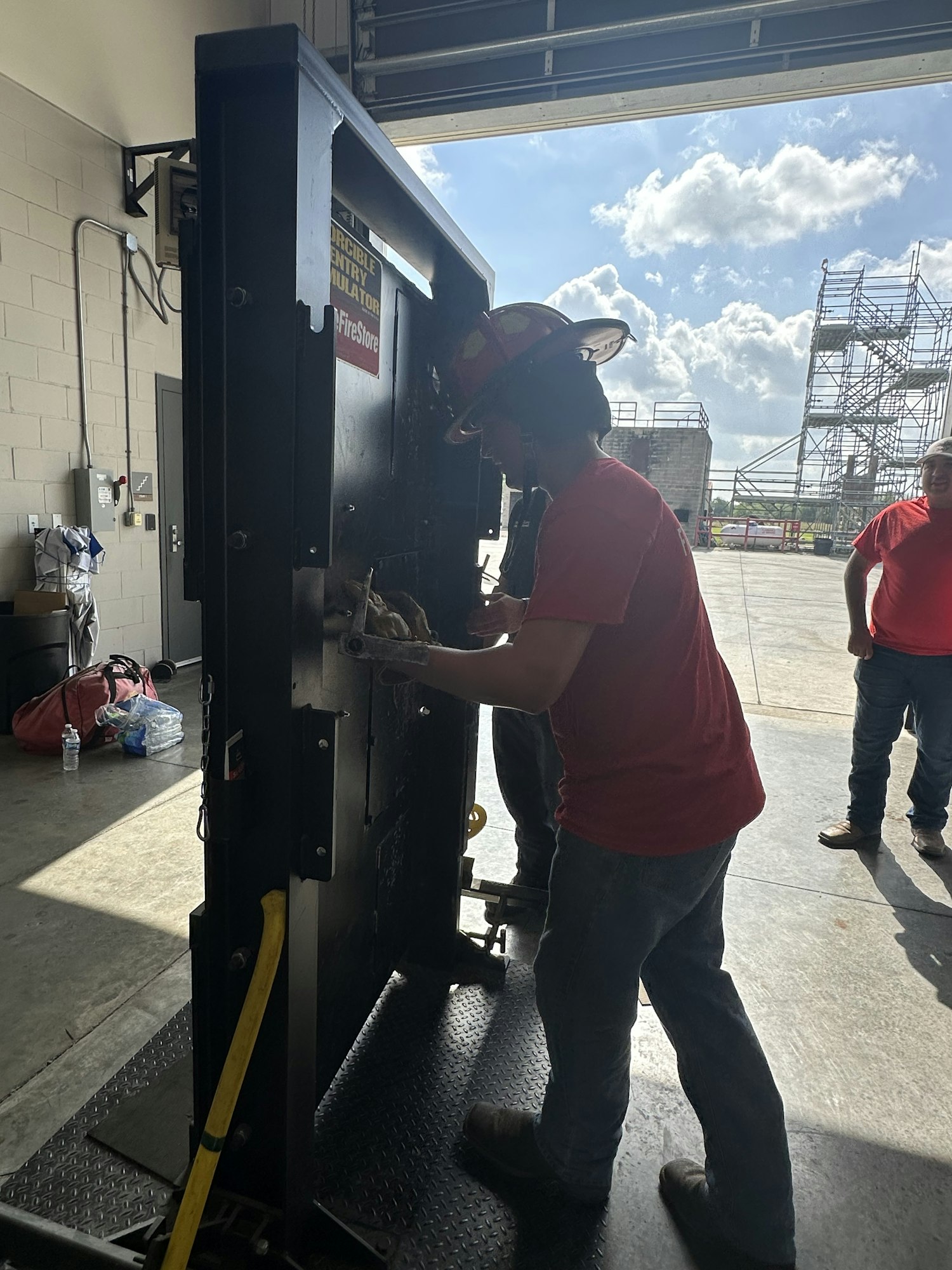 A person in a red shirt and helmet is working on machinery in a garage, with scaffolding visible outside.