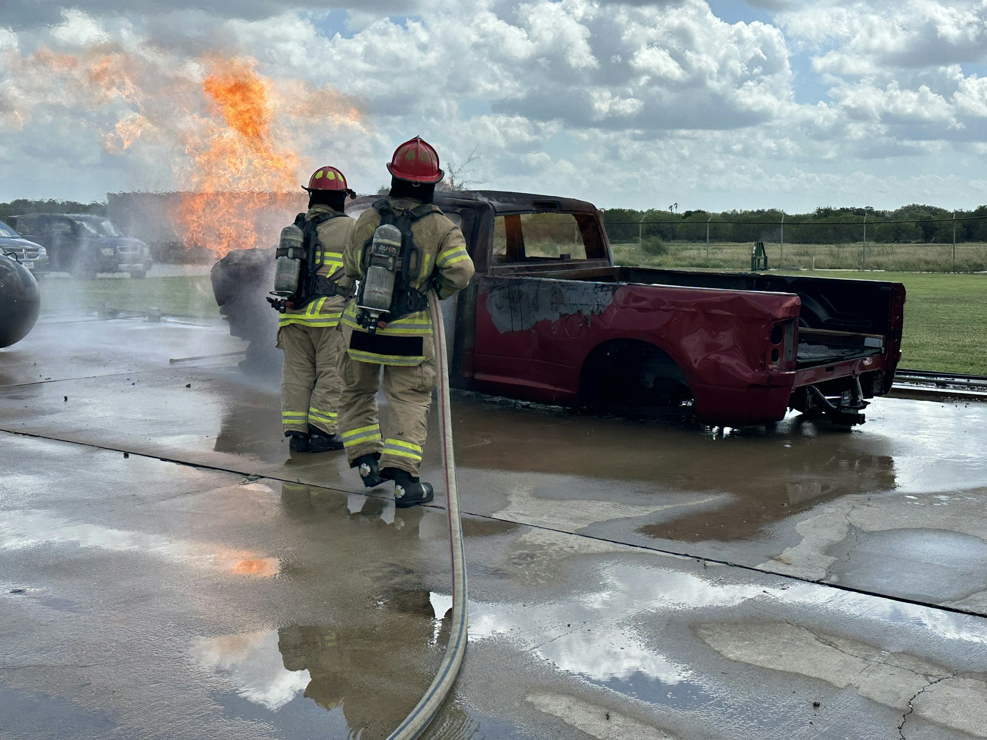 Firefighters extinguishing a fire on a red vehicle with flames in the background.
