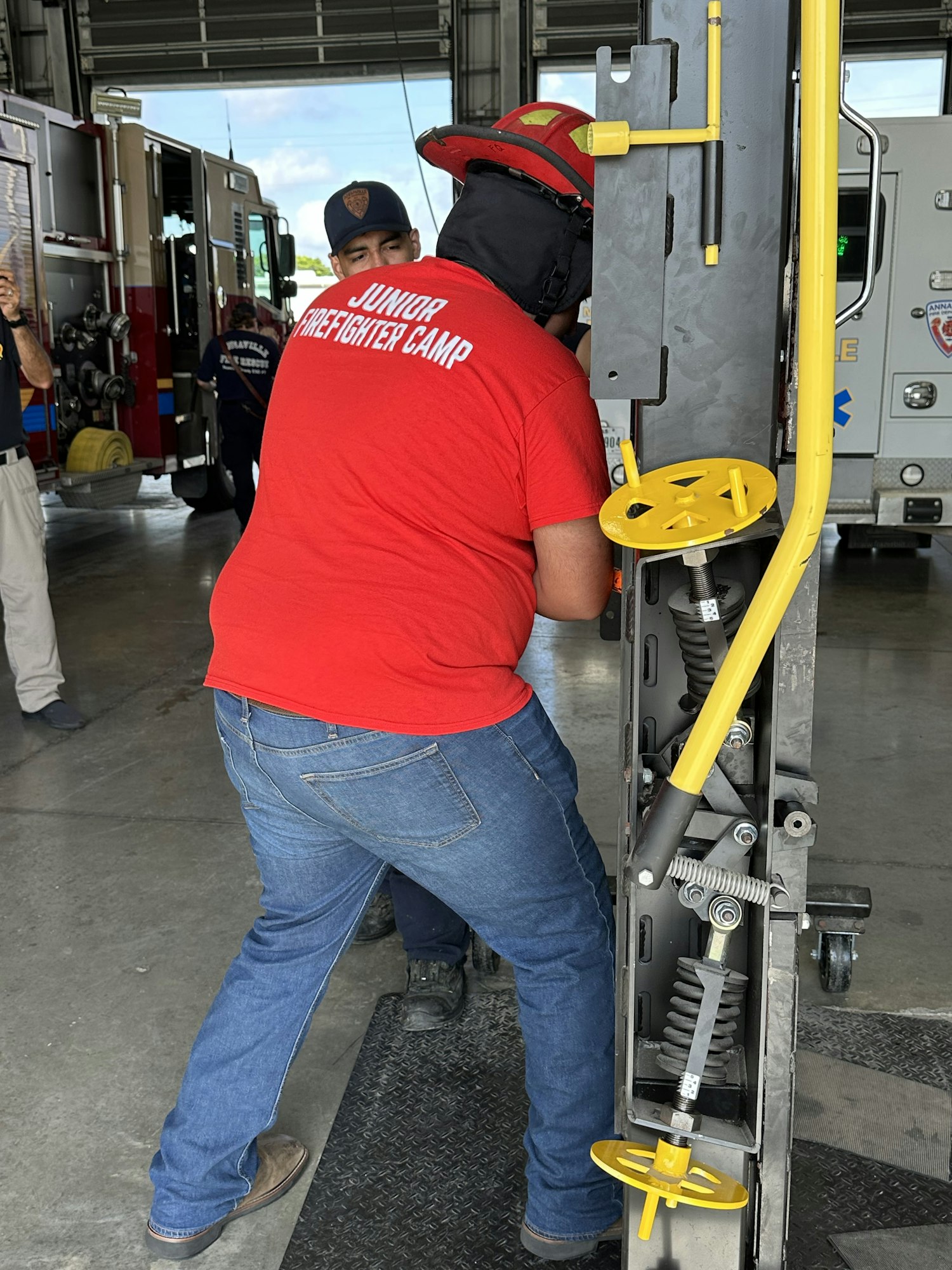 A person in a red shirt and cowboy hat stands at a metal apparatus in a garage near emergency vehicles.